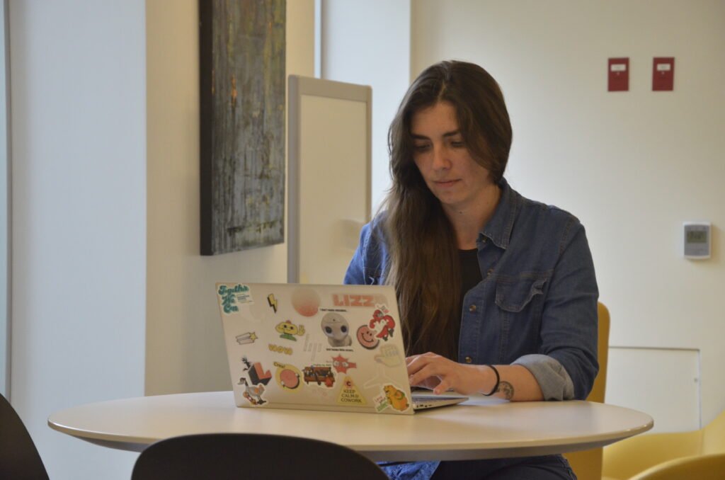 Deidre Bonilla, marketing strategist, working on a laptop with stickers while seated at a round table, wearing a denim-style blouse, with a white wall and framed picture in the background